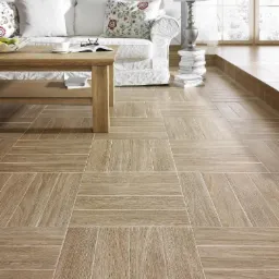 Jasna podłoga z paneli ułożonych w kwadraty, z widocznym fragmentem salonu z białym narożnikiem, drewnianym stolikiem i lampą na stosie książek.