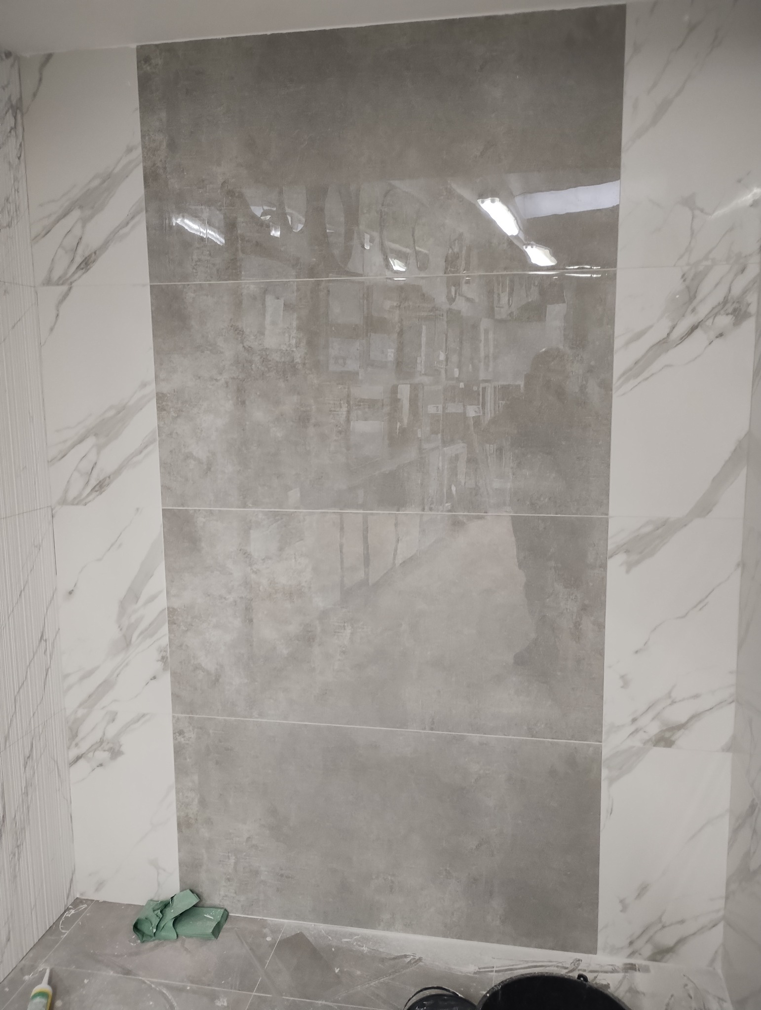 Ściana wyłożona szarymi i białymi płytkami imitującymi marmur, widoczne narzędzia i akcesoria podczas prac wykończeniowych w łazience.