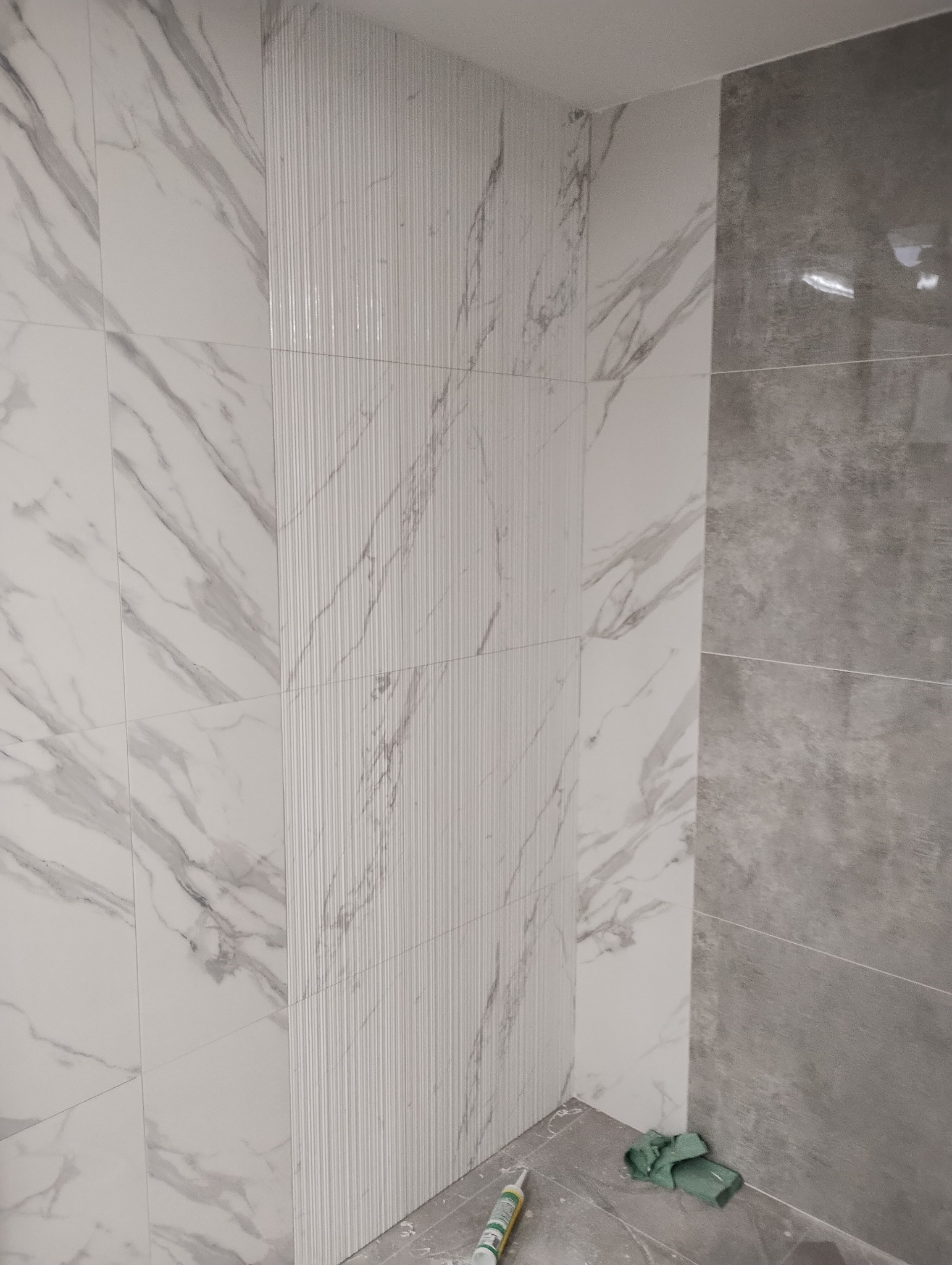 Fragment narożnika łazienki z ułożonymi płytkami: białe imitujące marmur z szarymi żyłami, białe z pionowymi żłobieniami oraz szare, widoczna tuba silikonu i ścierka na podłodze.