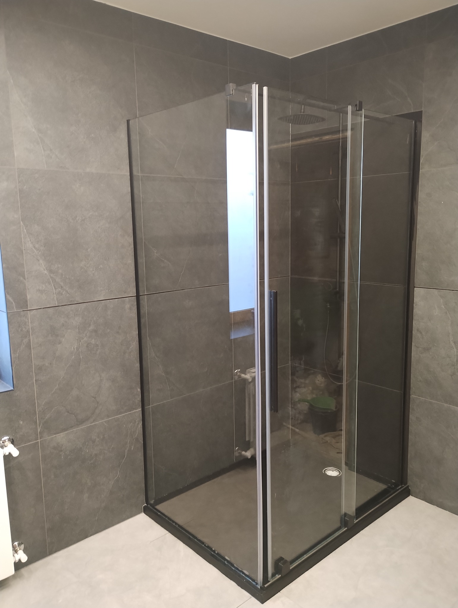 Nowoczesna kabina prysznicowa z czarnymi profilami i deszczownicą, szare płytki imitujące marmur na ścianach, widoczny grzejnik drabinkowy i odpływ liniowy.