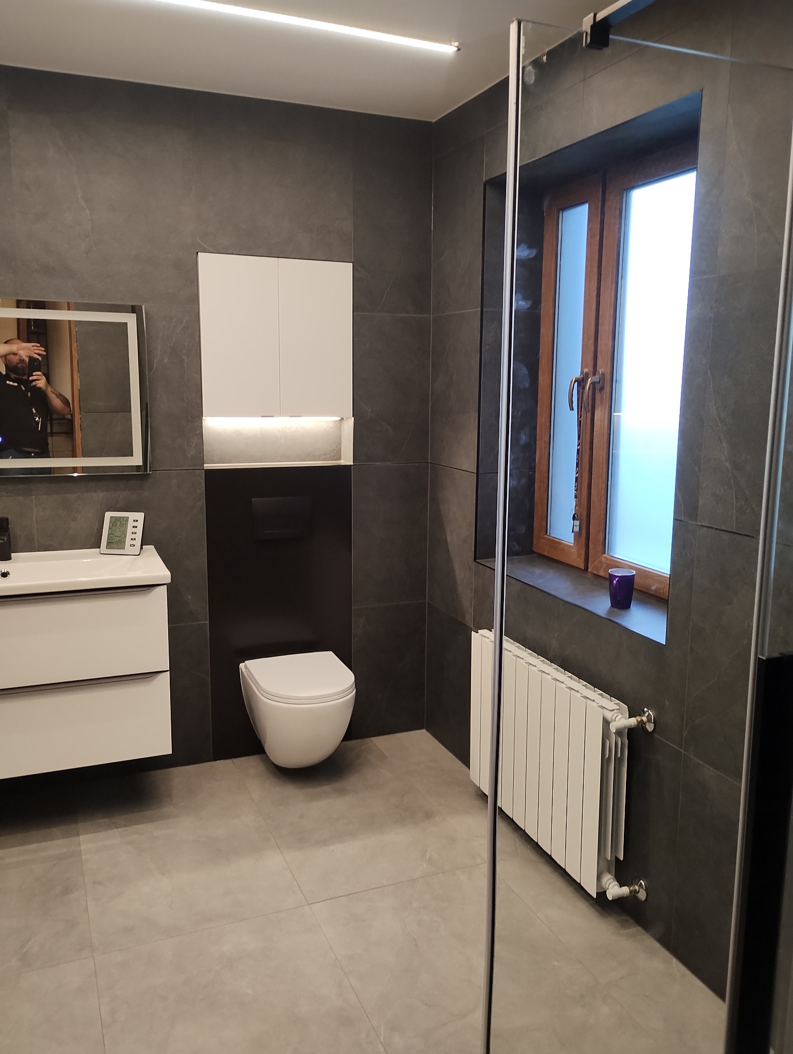 Nowoczesna łazienka z szarymi płytkami, wiszącą toaletą, białą szafką nad nią, szklaną kabiną prysznicową i grzejnikiem pod oknem z drewnianą ramą.