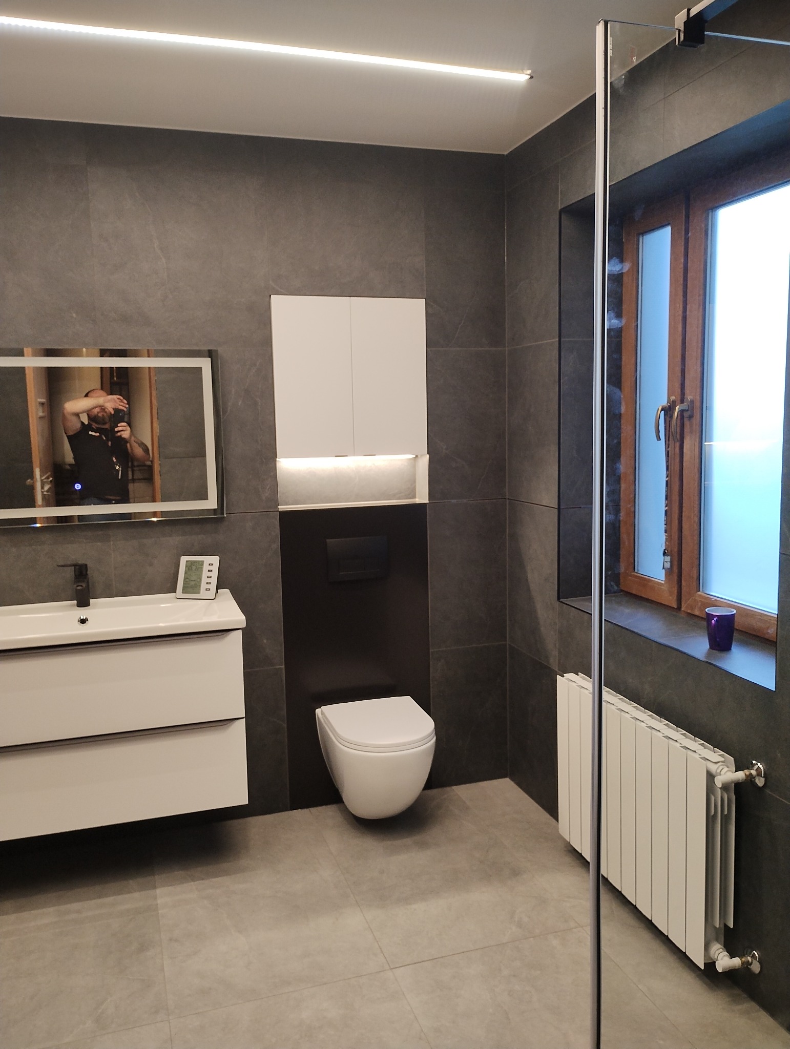 Nowoczesna łazienka z szarymi płytkami, podwieszaną toaletą, białą szafką nad toaletą z podświetleniem, umywalką z szafką i lustrem z podświetleniem, w którym odbija się osoba robiąca zdjęcie.