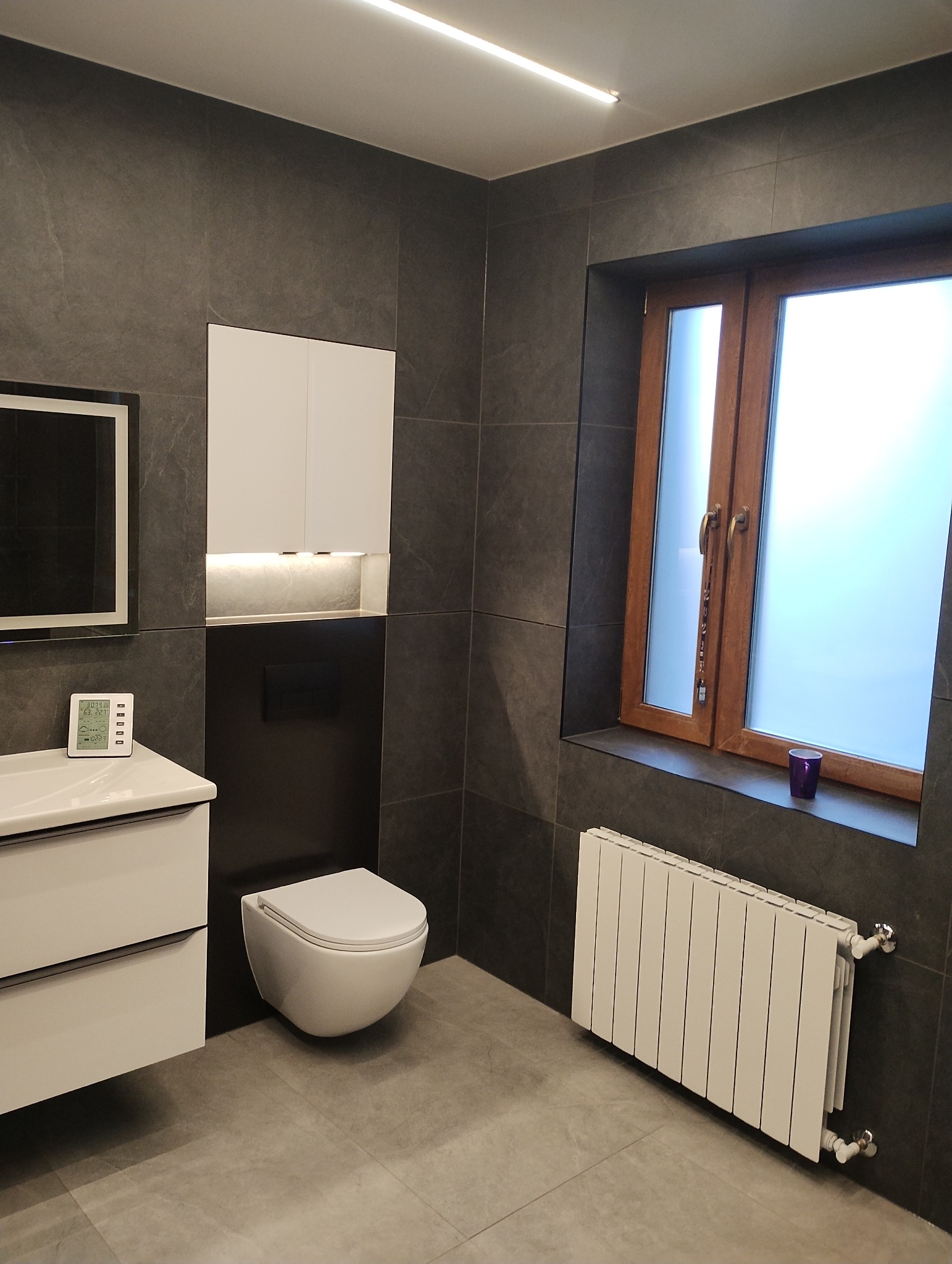 Nowoczesna łazienka z szarymi płytkami, podwieszaną toaletą, białymi szafkami i grzejnikiem pod oknem z drewnianą ramą, oświetlona listwą LED na suficie.
