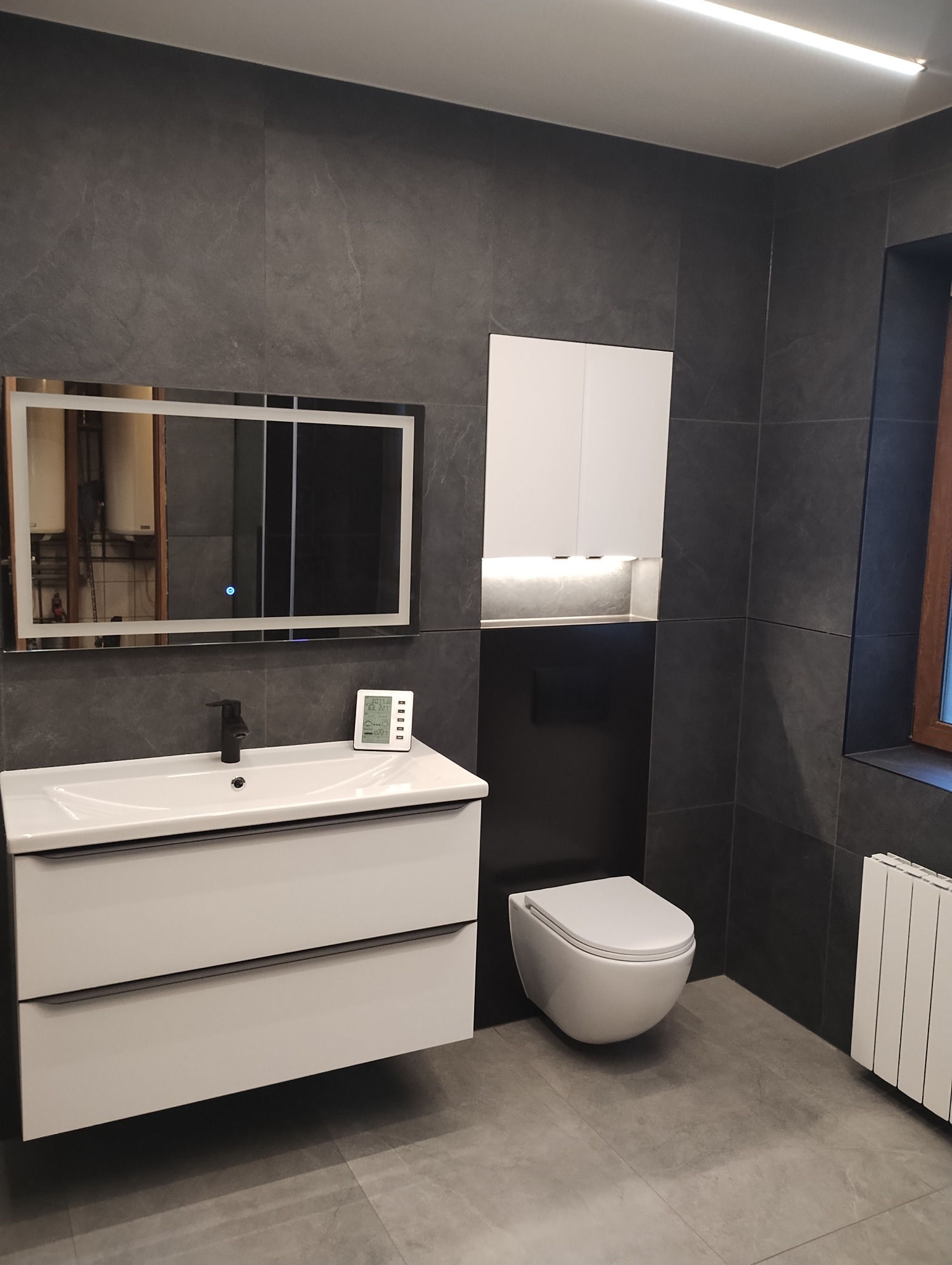 Nowoczesna łazienka w odcieniach szarości z białymi meblami: szafka pod umywalką z dwiema szufladami, wisząca toaleta, szafka wisząca z oświetleniem LED, lustro z podświetleniem LED i czarna armatura.