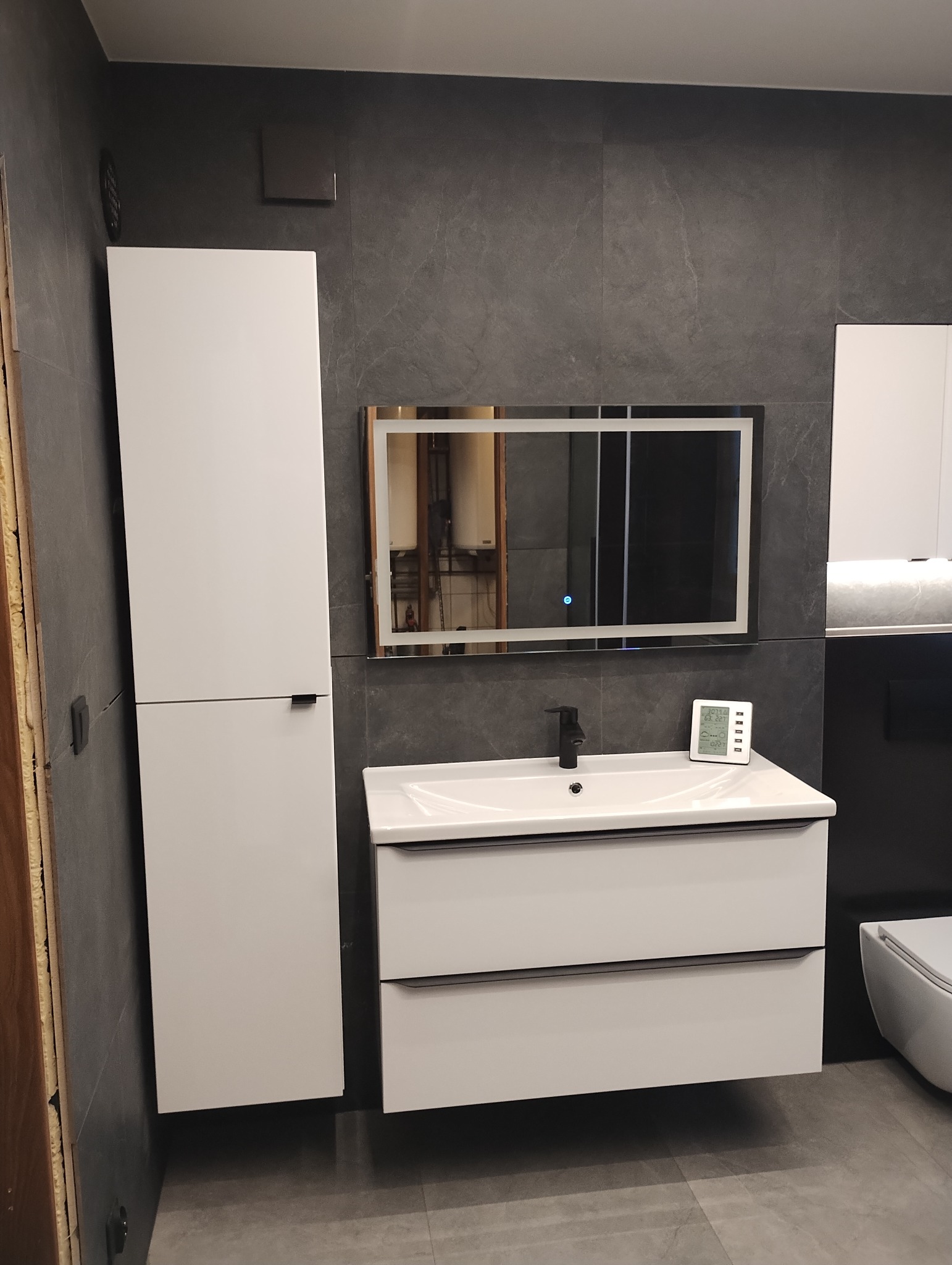Nowoczesna łazienka z szarymi płytkami, białymi szafkami pod umywalką i wysoką szafką, podświetlanym lustrem i czarną armaturą, uzupełniona o elektroniczny termometr.