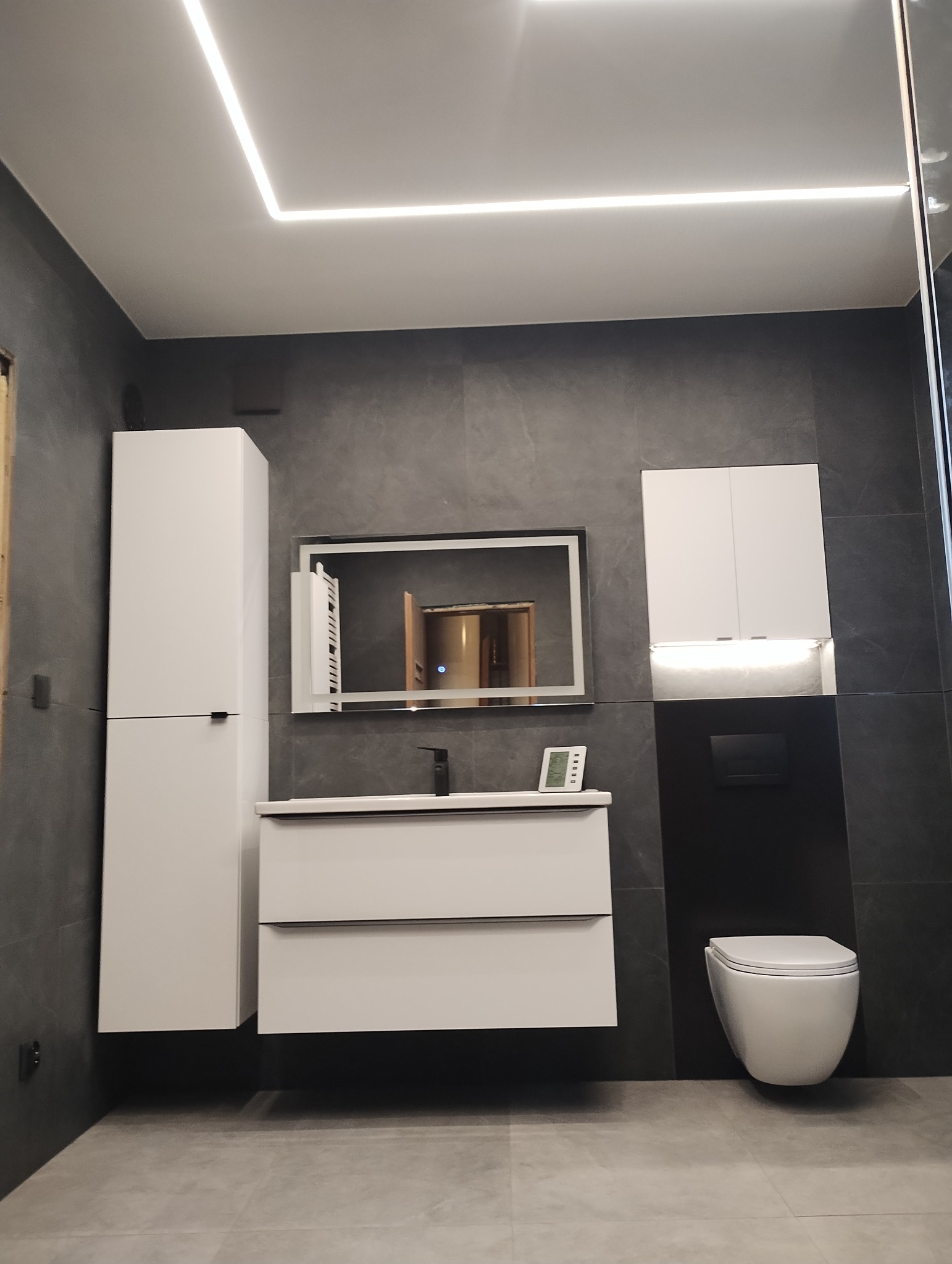Nowoczesna łazienka w szaro-białej kolorystyce z podwieszaną toaletą, umywalką z szafką, wysoką szafką i lustrem z podświetleniem LED, oświetlenie sufitowe w formie białych linii