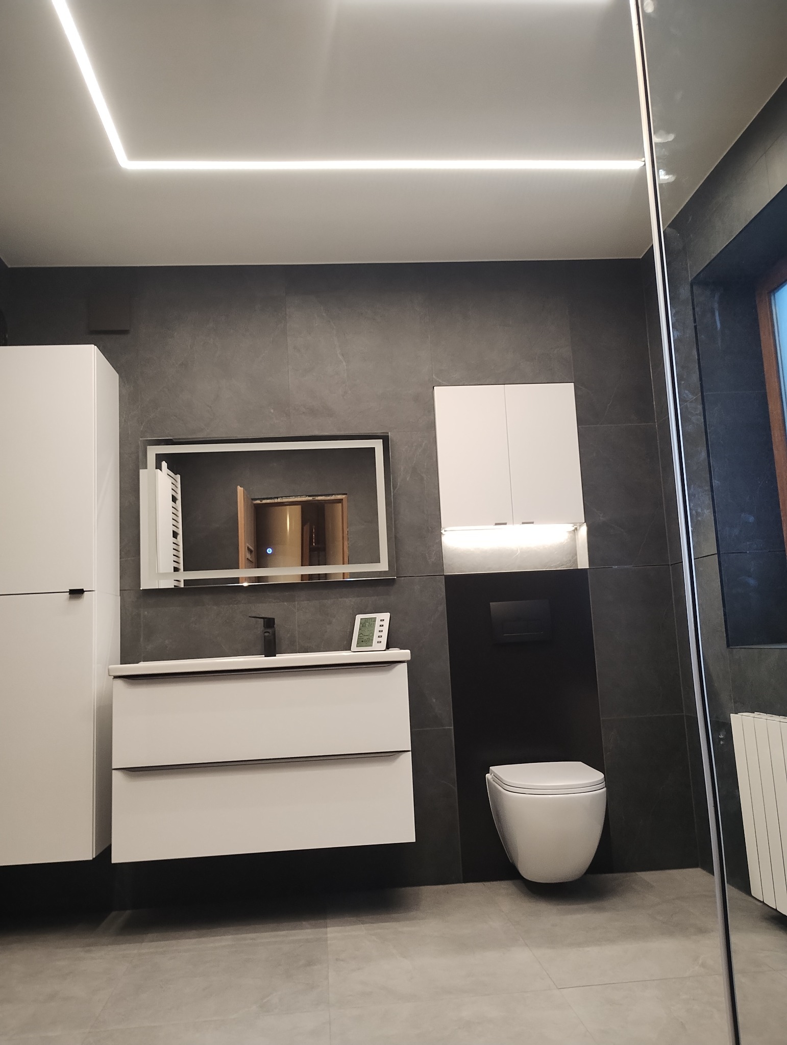 Nowoczesna łazienka z szarą płytką imitującą kamień, białymi szafkami wiszącymi i umywalką z szufladami, podświetlanym lustrem i podwieszaną toaletą. W lustrze odbija się fragment innego...