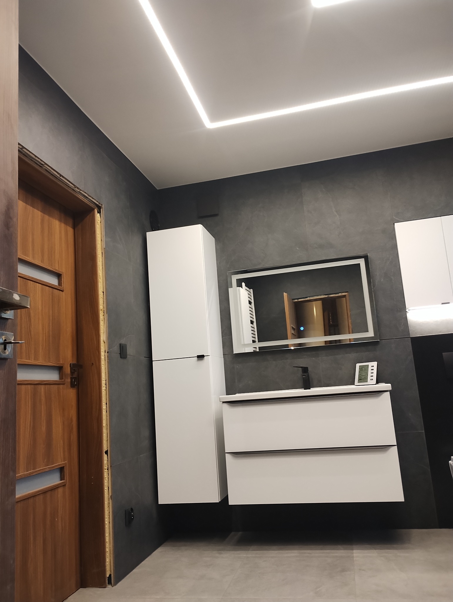 Nowoczesna łazienka z szarą płytką, białymi szafkami pod umywalką i wysoką szafką wiszącą, lustro z podświetleniem LED, oraz podświetlenie sufitowe w formie kwadratu, widoczna drewniana ościeżnica...