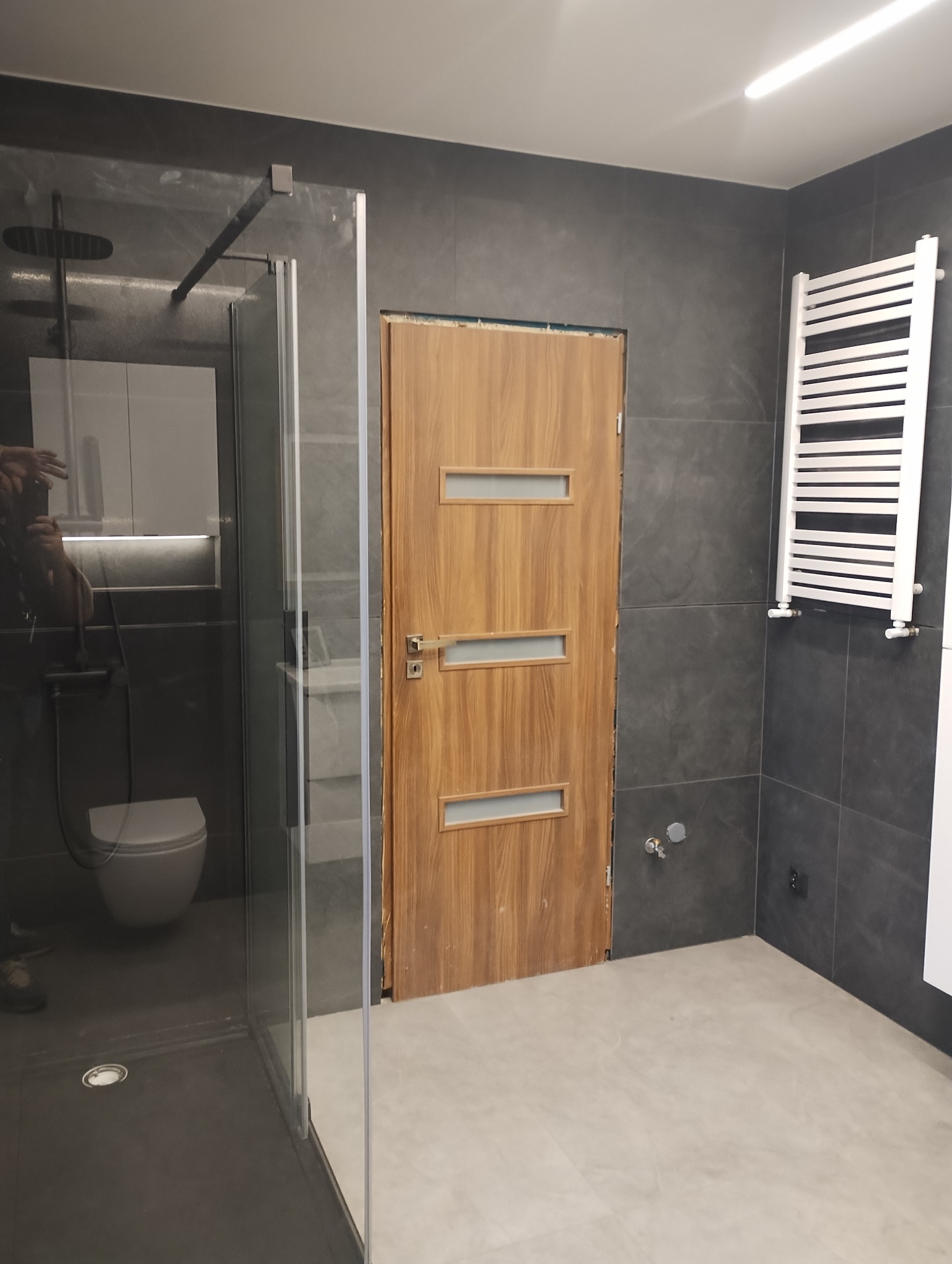 Nowoczesna łazienka z kabiną prysznicową typu walk-in, drewnianymi drzwiami z przeszkleniami i grzejnikiem drabinkowym na ścianie z szarych płytek.