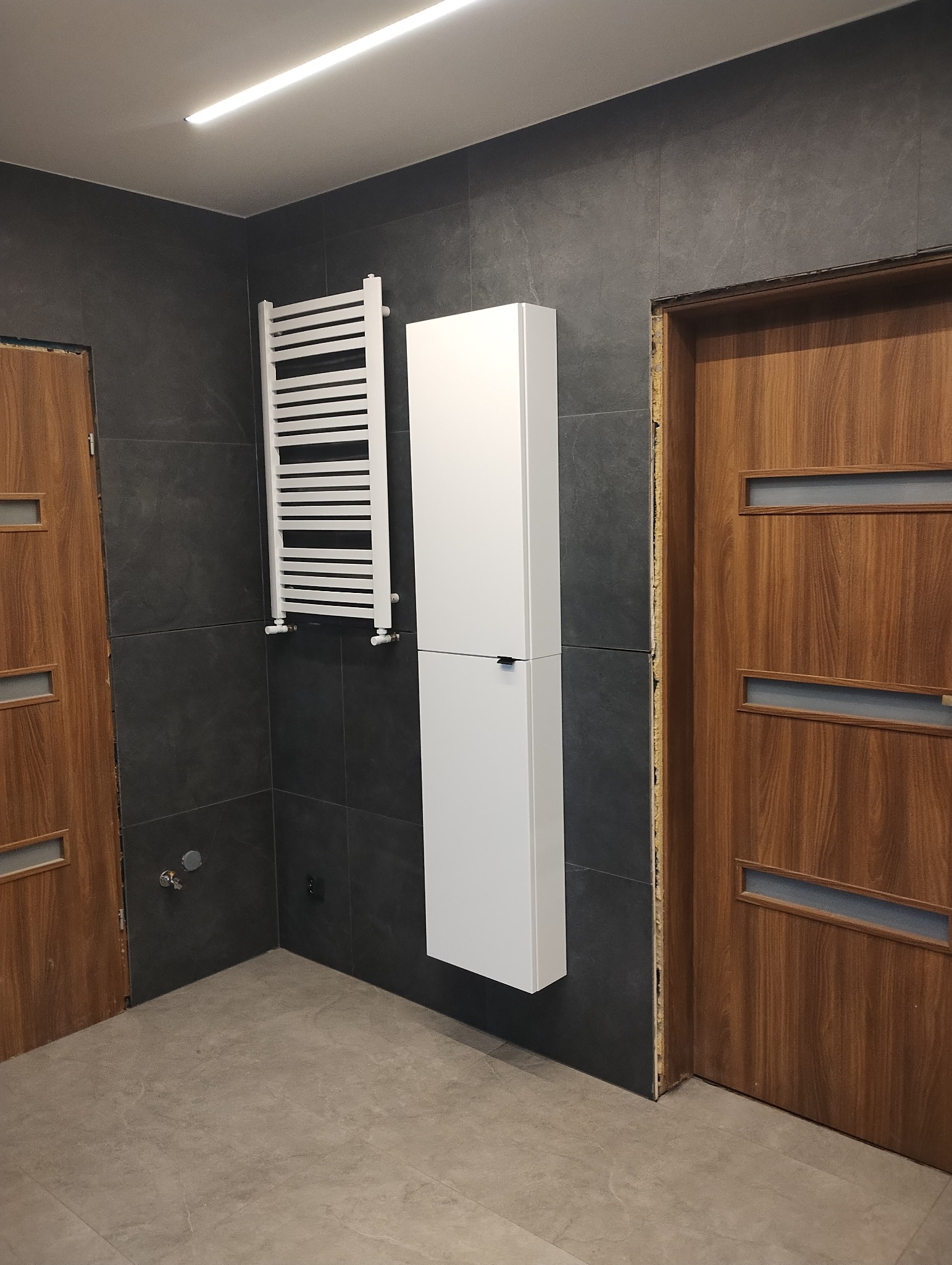 Nowoczesna łazienka z ciemnymi płytkami, białym grzejnikiem drabinkowym i wysoką szafką wiszącą; widoczne niedokończone obramowania drzwi.