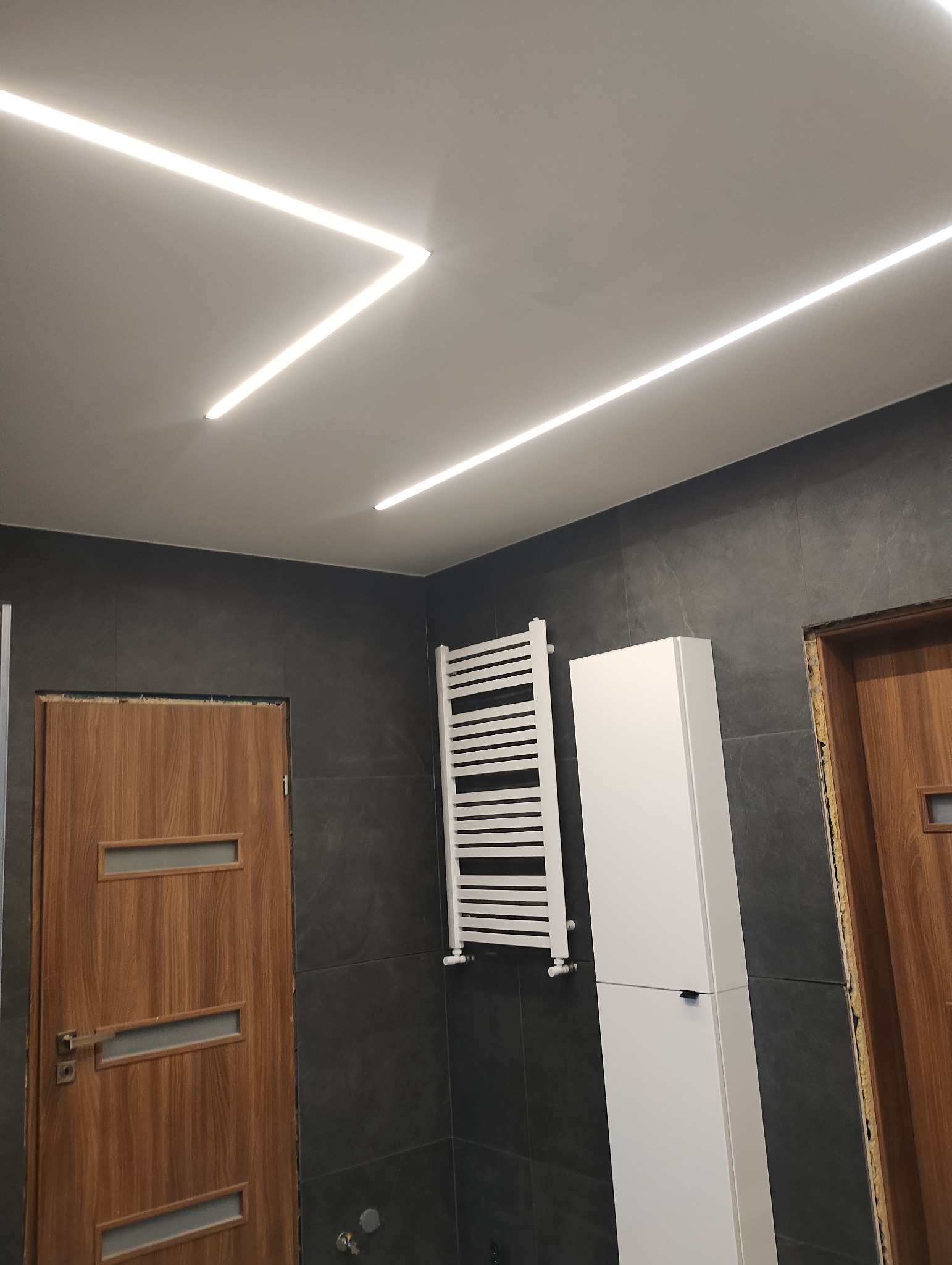Nowoczesna łazienka z szarymi płytkami, podświetlanym sufitem LED, grzejnikiem drabinkowym i drewnianymi drzwiami z przeszkleniami.