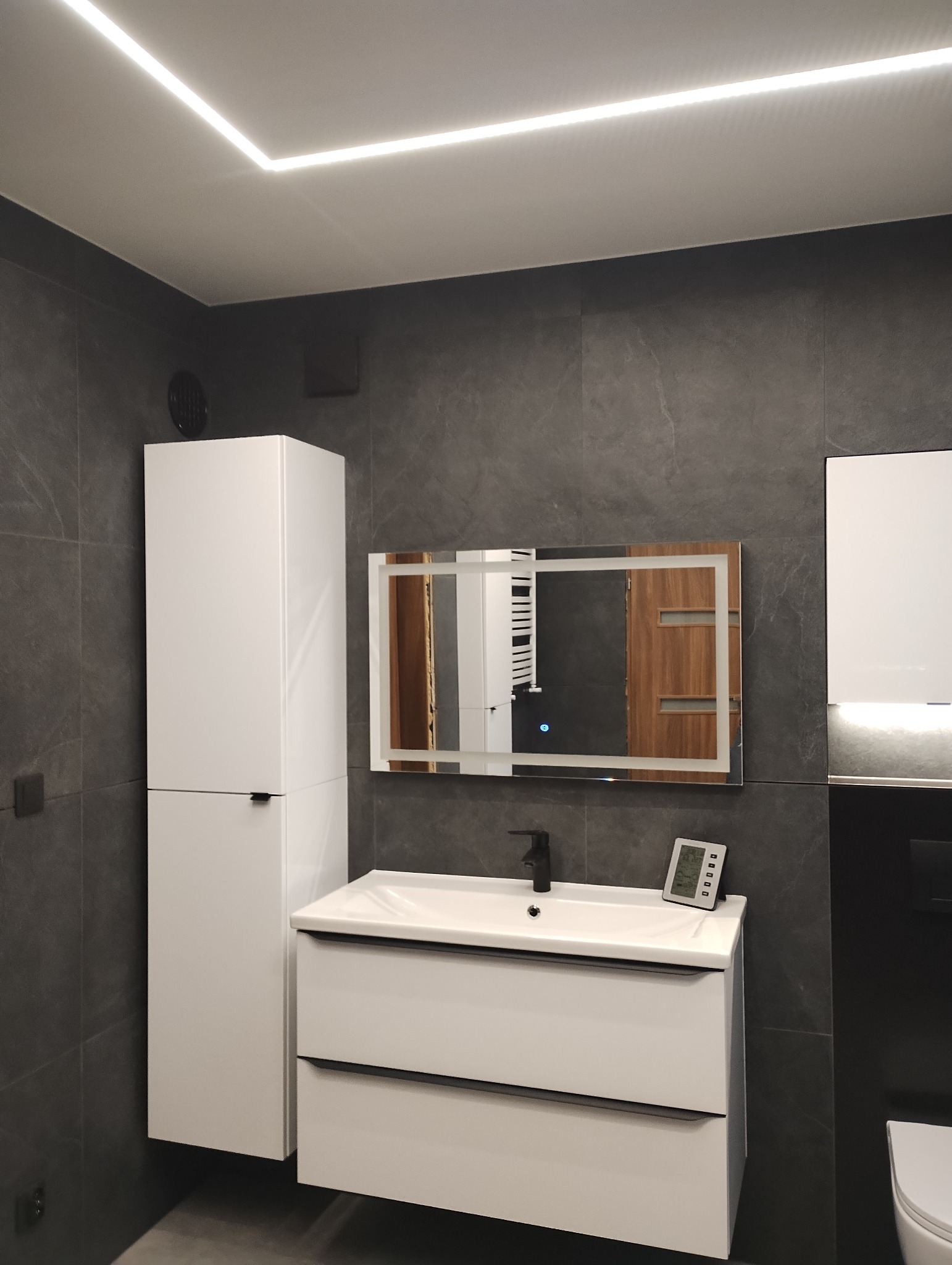 Nowoczesna łazienka z szarymi płytkami, białymi szafkami pod umywalką i wysoką szafką, podświetlanym lustrem oraz czarną armaturą i termometrem na blacie.