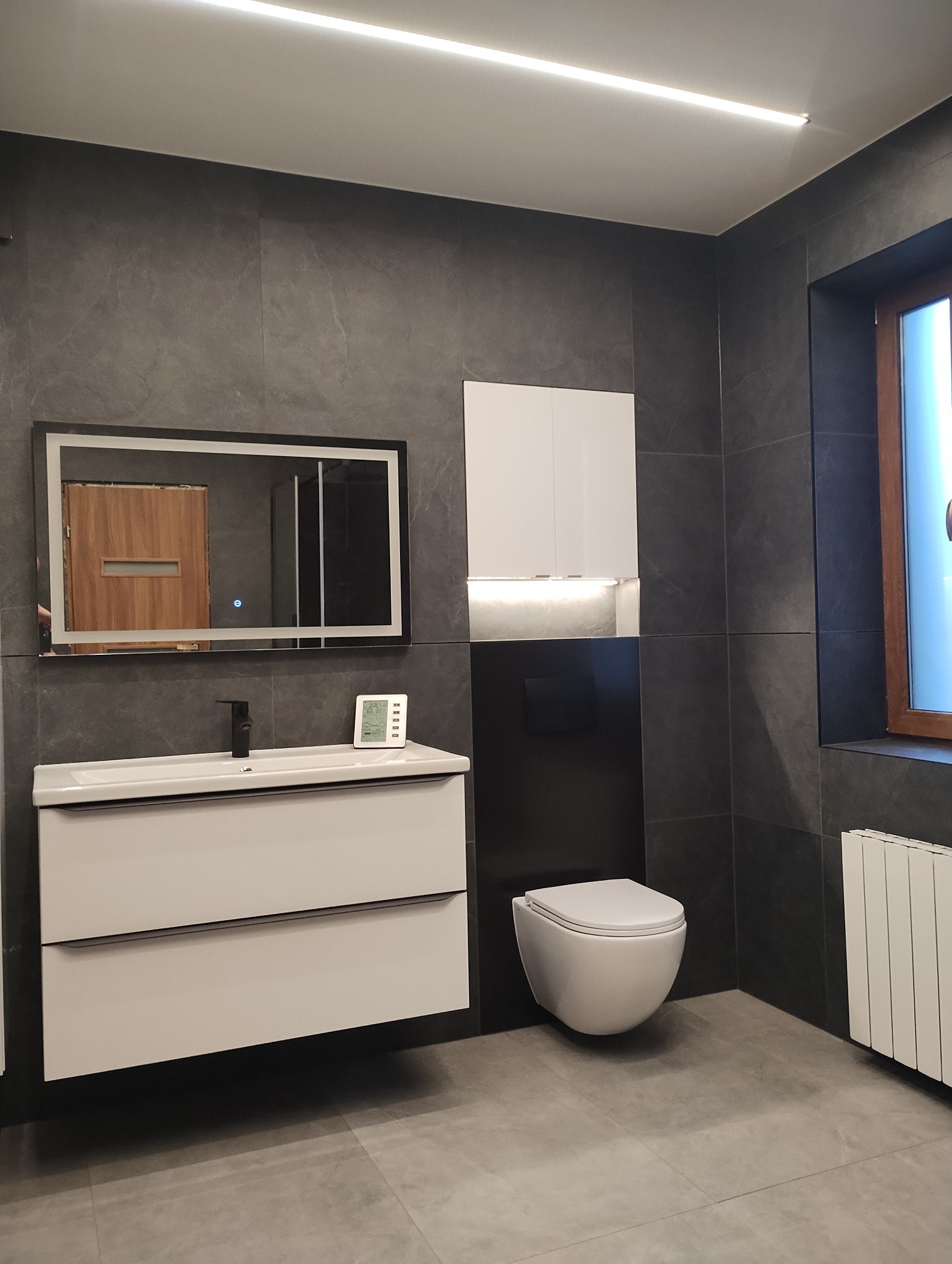 Nowoczesna łazienka z szarymi płytkami, białymi meblami łazienkowymi, podświetlanym lustrem i wiszącą toaletą, oświetlona ledowym światłem sufitowym.