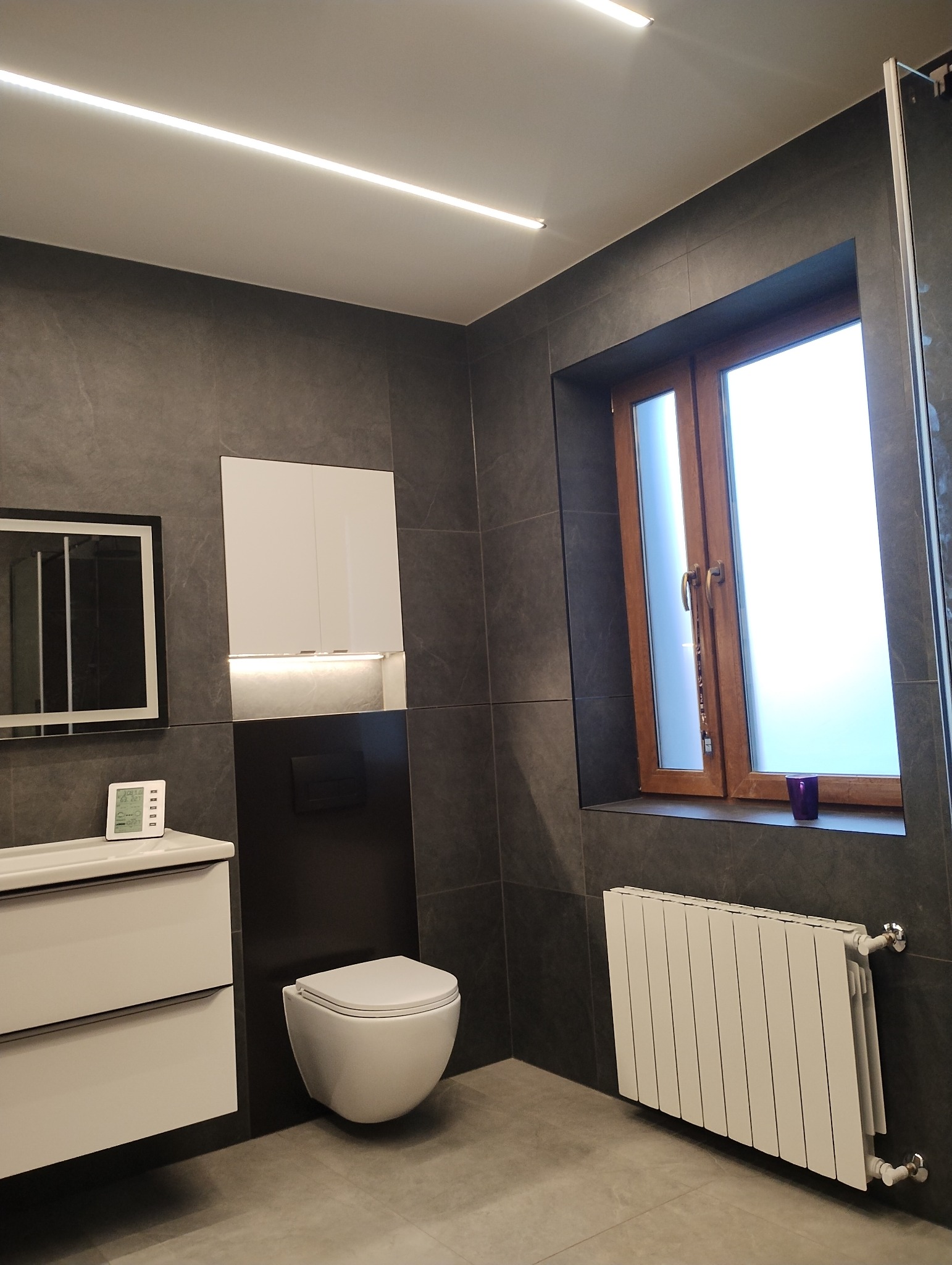 Nowoczesna łazienka z szarymi płytkami, podwieszaną toaletą, białą szafką z lustrem i drewnianą ramą okienną z fioletowym wazonem na parapecie, oświetlona liniowymi lampami LED na suficie.