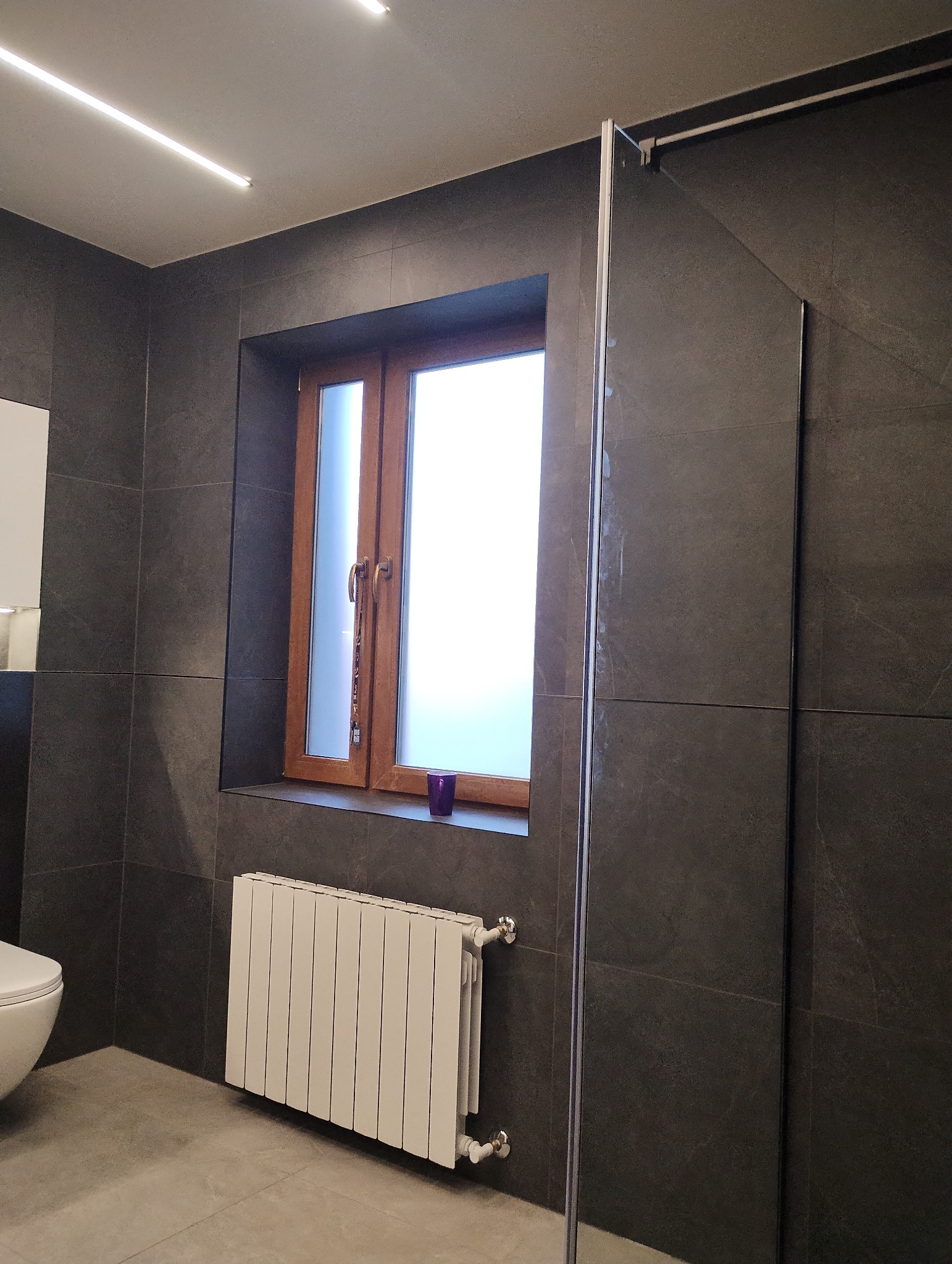 Szara łazienka z drewnianym oknem, białym grzejnikiem i szklaną kabiną prysznicową, gdzie na parapecie stoi mały fioletowy wazonik.