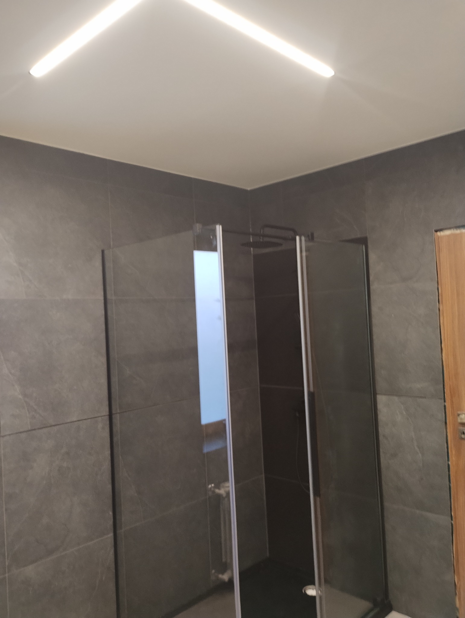 Kabina prysznicowa z grafitowymi płytkami w nowoczesnej łazience, oświetlona podłużną lampą sufitową, widok na fragment drzwi.