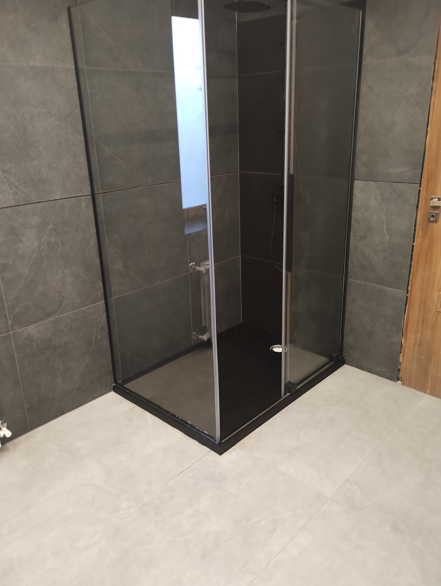 Nowoczesna kabina prysznicowa z ciemnymi płytkami na ścianach i podłodze, widok z niskiego kąta, drzwi szklane z czarnymi ramami, jasna podłoga w łazience.