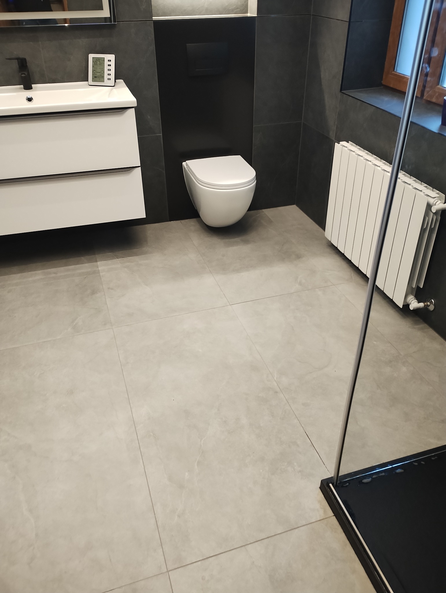 Nowoczesna łazienka z szarymi płytkami podłogowymi i ściennymi, białą wiszącą toaletą oraz umywalką z szafką, widoczny grzejnik i fragment kabiny prysznicowej z czarnym brodzikiem.