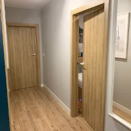 Wnętrze mieszkania z dwiema drewnopodobnymi ościeżnicami drzwiowymi, jedna zamknięta, druga otwarta, ukazująca fragment sypialni, oraz lustro w białej ramie z odbiciem podłogi.