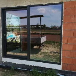 Duże przesuwne okno z ciemną ramą w surowym stanie budynku, widok na podwórze z odbijającym się na szybie wózkiem transportowym.