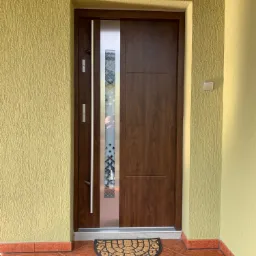 Brązowe drzwi wejściowe z przeszkleniem w geometryczny wzór, stalowy uchwyt, wycieraczka przed wejściem, ściana o fakturze baranka w odcieniach żółci.