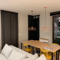 Nowoczesne wnętrze z ciemnymi, drewnianymi żaluzjami poziomymi w oknach, oświetlone wiszącymi lampami z miedzianymi kloszami nad drewnianym stołem z kolorowymi krzesłami.