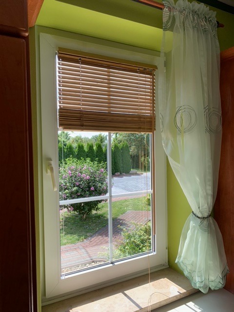 Białe okno z drewnianymi żaluzjami wewnętrznymi i zwiewną, jasną firanką, widok na zadbany ogród.