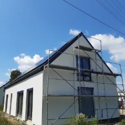 Nowoczesny dom jednorodzinny z białą elewacją i ciemną stolarką okienną, w trakcie prac wykończeniowych, widoczne rusztowania i zabezpieczone okna folią, błękitne niebo z chmurami w tle.