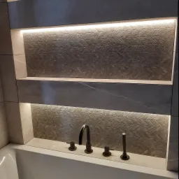 Nowoczesna łazienka z podświetlanymi wnękami wyłożonymi płytkami o wzorze jodełki, bateria umywalkowa w kolorze ciemnego brązu, biała wanna.