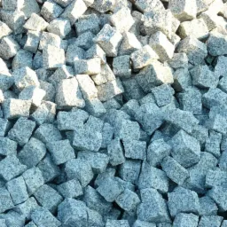 Stos szarych, surowych kostek o nieregularnych kształtach, oświetlonych z góry, z widoczną teksturą kamienia.