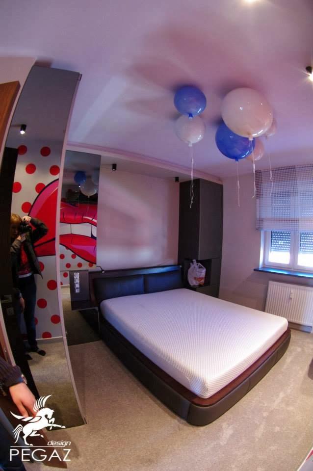 Nowoczesna sypialnia z czarną ramą łóżka, białym materacem, balonami pod sufitem i lustrem z motywem ust w stylu pop-art na ścianie.