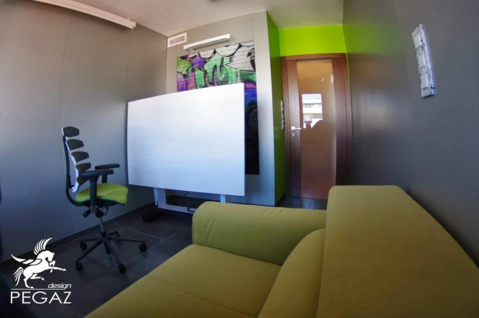 Biuro z zielonymi akcentami: sofa, krzesło, ściana obok brązowych drzwi, biała tablica na tle graffiti, szare ściany i podłoga.