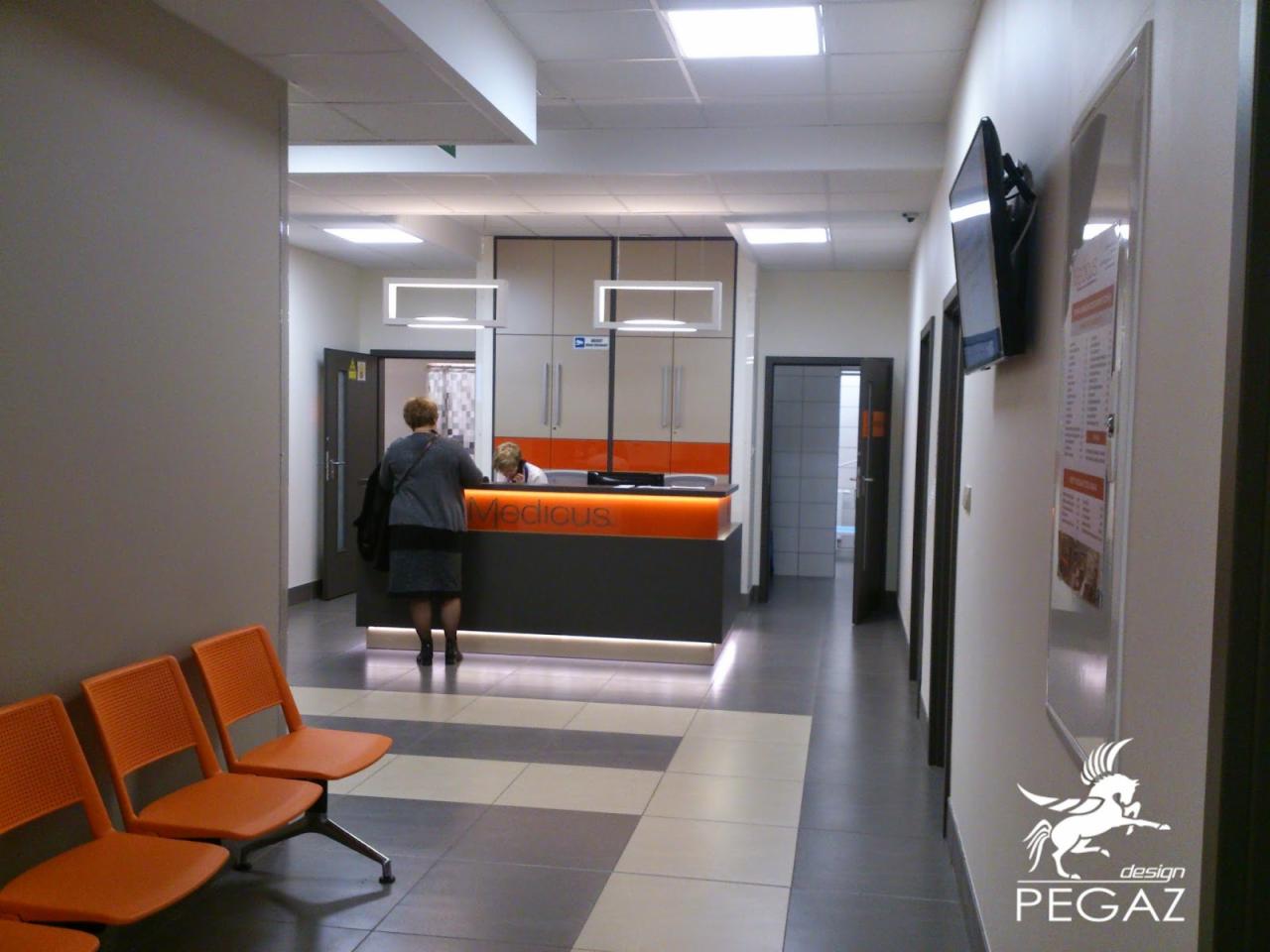 Korytarz w placówce medycznej z recepcją podświetloną pomarańczowym światłem, pomarańczowe krzesła dla pacjentów, tablica informacyjna na ścianie i logo firmy projektowej w prawym dolnym rogu.