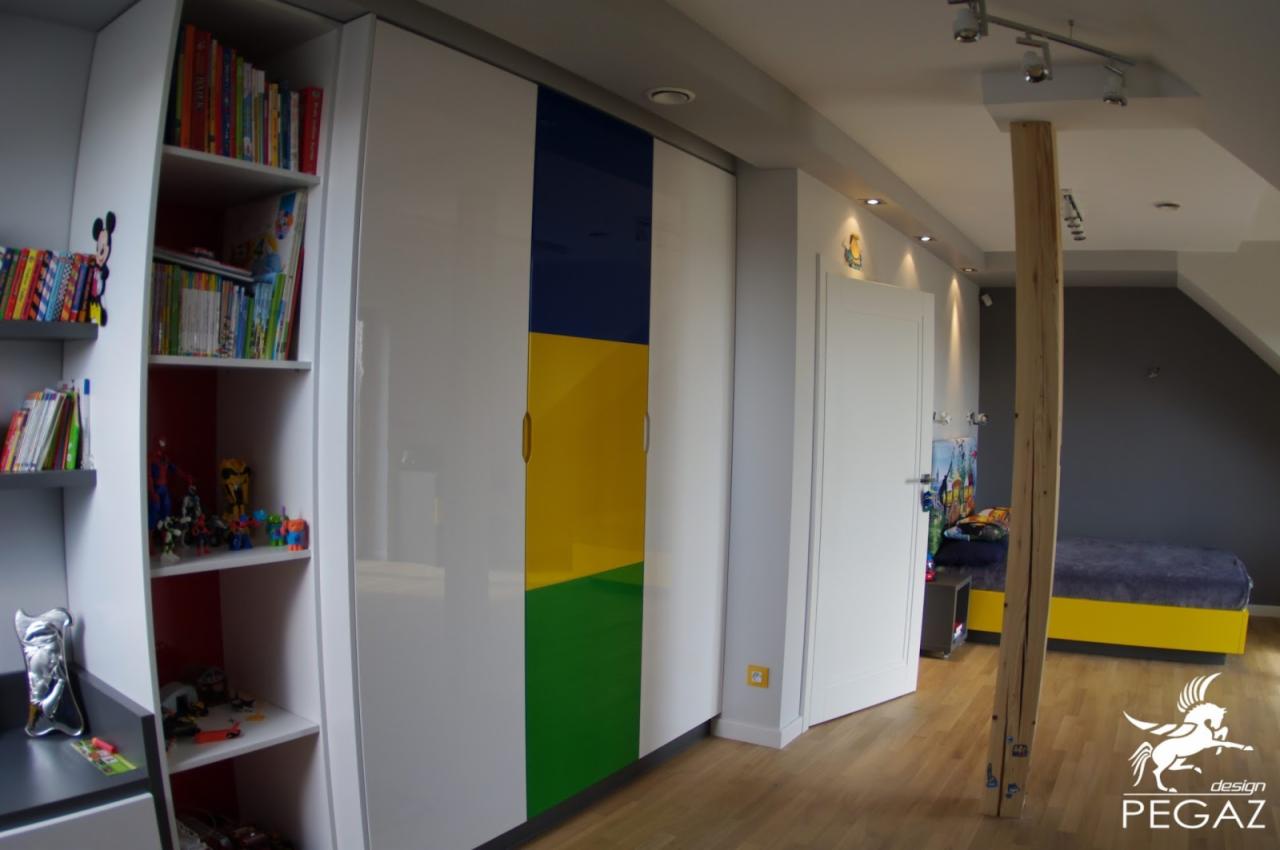 Pokój dziecięcy z białymi szafami z kolorowymi panelami (niebieski, żółty, zielony), otwartym regałem z książkami i zabawkami, oraz łóżkiem z żółtą podstawą w tle.