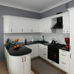 Klasyczna kuchnia z białymi szafkami, czarnym blatem i mozaiką na ścianie, uzupełniona czajnikiem, ekspresem do kawy i dywanem w orientalny wzór.