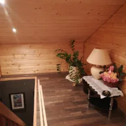 Drewniane wnętrze poddasza z widocznymi panelami podłogowymi i boazerią, oświetlone lampą stojącą. Widoczne listwy przypodłogowe przed montażem.