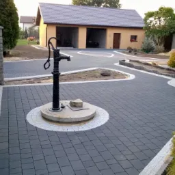 Ułożona szara kostka brukowa z dekoracyjnym obramowaniem i studnią z pompą na posesji. Widoczny budynek gospodarczy z dwoma bramami garażowymi w tle.