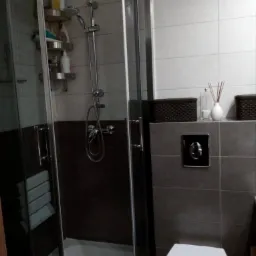 Wykończona łazienka z kabiną prysznicową z hydromasażem, półkami na kosmetyki i siedziskiem, obok toaleta i szare płytki z dekoracyjnymi koszykami i dyfuzorem zapachowym.