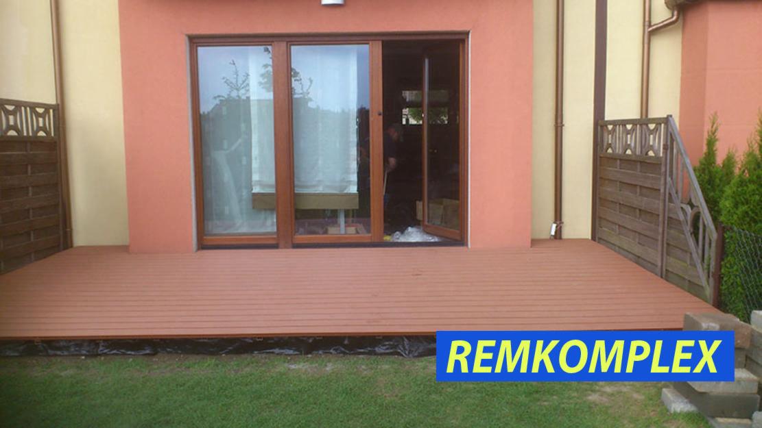 Brązowy taras z desek kompozytowych przed domem z pomarańczową elewacją, widoczne przesuwne drzwi balkonowe i fragment ogrodu z kostką brukową oraz logo firmy REMKOMPLEX.