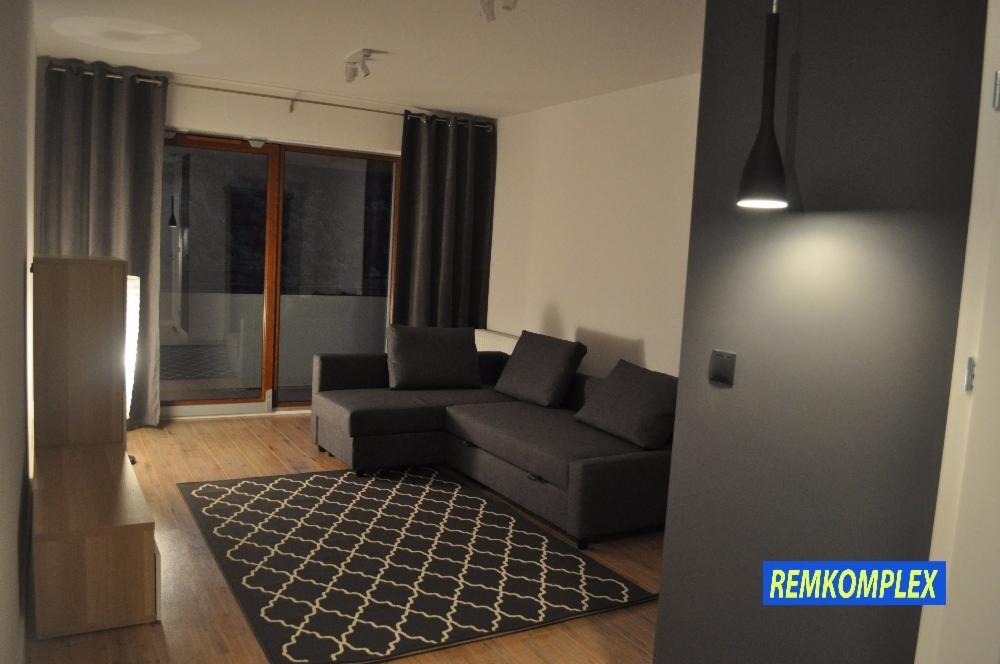 Nowoczesny salon z szarą narożną sofą, wzorzystym dywanem, drewnianą podłogą i minimalistyczną lampą wiszącą, widziany z perspektywy wejścia.