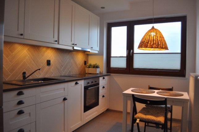 Jasna kuchnia z drewnianą mozaiką na ścianie, białymi szafkami z czarnymi uchwytami, okapem, oknem z roletami plisowanymi i lampą z wikliny nad stołem.