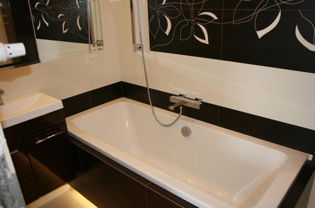 Biała wanna z chromowaną armaturą i słuchawką prysznicową na tle czarno-białych płytek z motywem kwiatowym. Umywalka z szafką w kolorze ciemnego drewna.