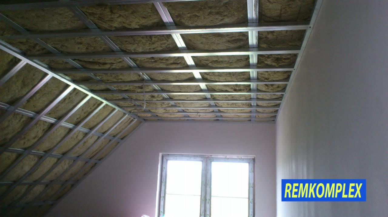 Montaż izolacji z wełny mineralnej w metalowym stelażu sufitu podwieszanego na poddaszu, widoczne okno i logo firmy.