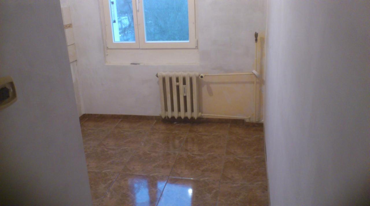 Wnętrze pokoju z brązową podłogą z płytek, białymi ścianami, grzejnikiem pod oknem i fragmentem drzwi po lewej stronie.