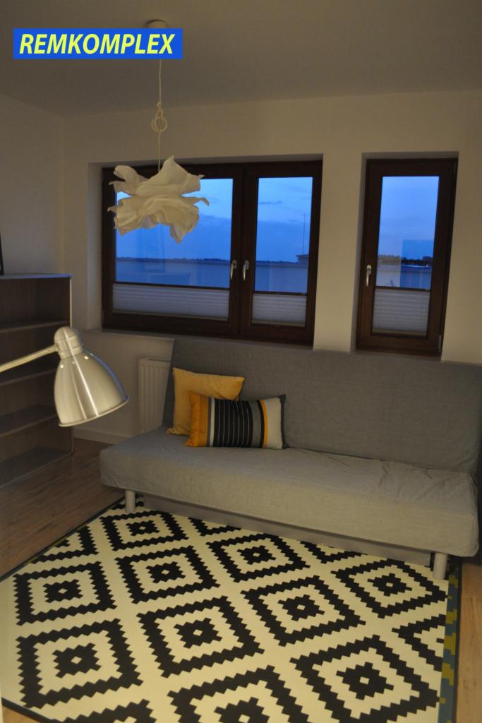 Jasne wnętrze pokoju z szarą sofą, wzorzystym dywanem w czarno-białe romby, lampą podłogową i oknami z widokiem na nocne niebo. Na suficie nietypowa lampa z papieru. Widoczny znak wodny w lewym...