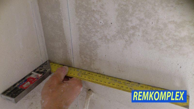 Pomiar kąta prostego w rogu pomieszczenia za pomocą kątownika i miarki. Ściany z surowym wykończeniem, widoczne ślady po szpachlowaniu.