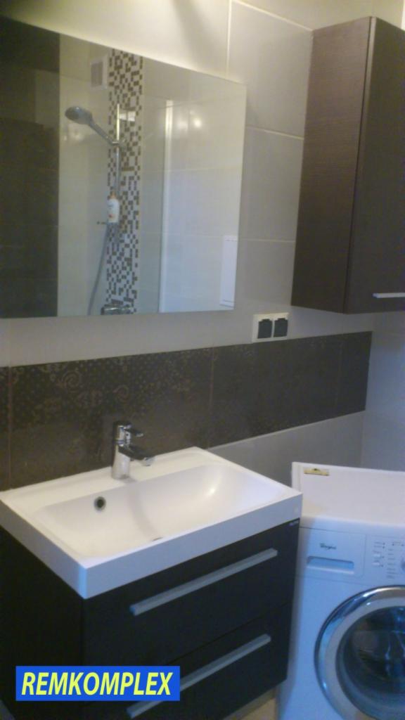 Wykończona łazienka z umywalką na szafce, lustrem, mozaiką pod prysznicem i pralką. Ciemne płytki dekoracyjne. Widoczny fragment kabiny prysznicowej.