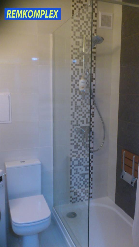Wykończona łazienka z kabiną prysznicową za szkłem, mozaiką na ścianie i toaletą. Widoczne płytki w jasnych odcieniach i element wentylacji.