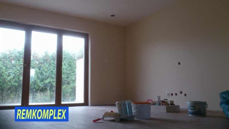 Wnętrze pokoju w trakcie malowania: ściany w beżowym odcieniu, duże okno z widokiem na zieleń, akcesoria malarskie rozłożone na podłodze. Przygotowanie do malowania ścian.