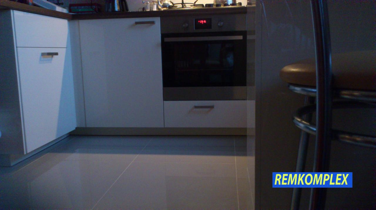 Nowoczesna zabudowa kuchenna w jasnych barwach z widocznym piekarnikiem i błyszczącą podłogą. Fragment krzesła barowego w tle.